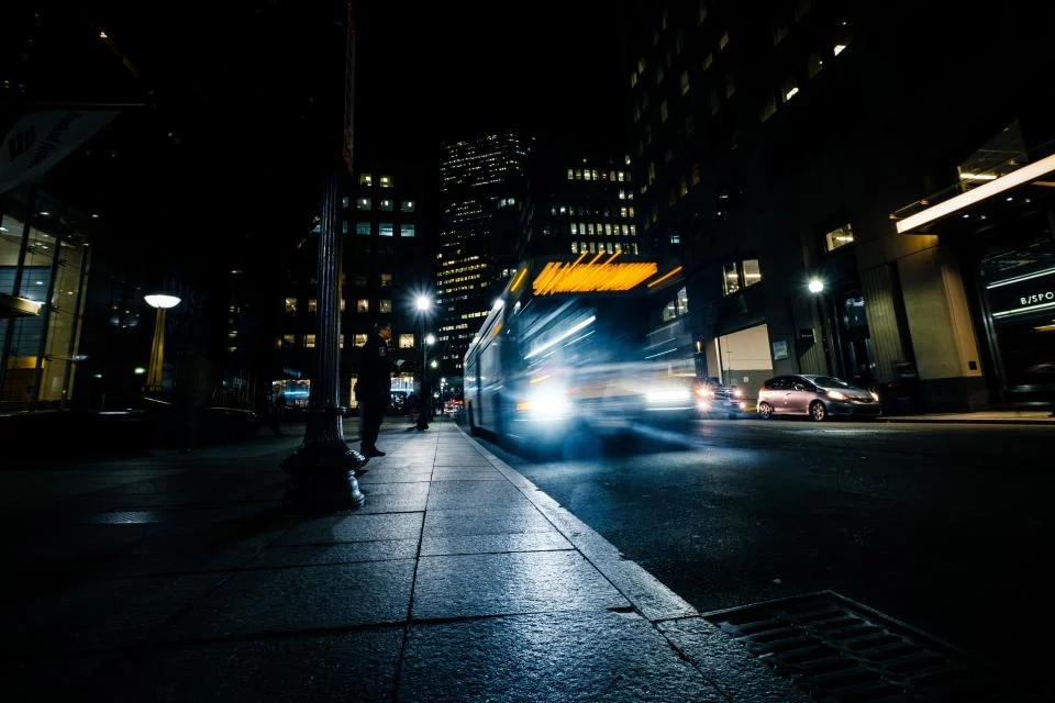 A bus pulling away from a stop on a dark city street, its lights blurred by a long exposure
